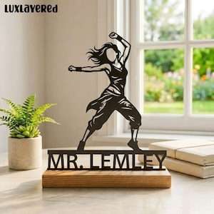 May include: A black metal sculpture of a person in a dance pose, mounted on a wooden base. The sculpture is of a person with arms raised, and the name "MR. LEMLEY" is displayed on the base. The sculpture is set on a table near a window.