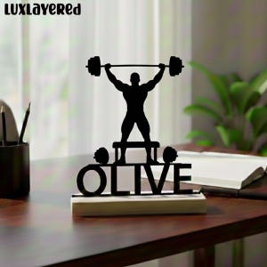 Pode incluir: Silhueta preta de um halterofilista levantando uma barra, com o nome "OLIVE" abaixo. A figura está sobre uma plataforma com pesos em ambos os lados. O design é montado em uma base de madeira.