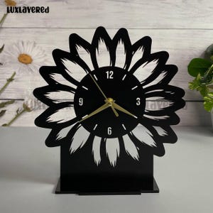 May include: A black sunflower-shaped desk clock with gold-colored hands. The clock face displays the numbers 12, 3, 6, and 9. The clock is made of a solid material and sits on a rectangular base.