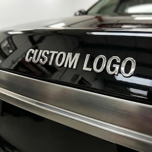Könnte beinhalten: Eine glänzend schwarze Fahrzeugoberfläche mit dem silbernen, metallischen Schriftzug CUSTOM LOGO in einer serifenlosen Schriftart. Unter dem Text verläuft eine horizontale Zierleiste aus gebürstetem Metall über die Breite des Paneels.