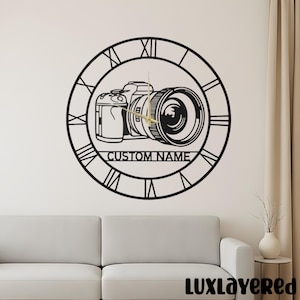 Puede incluir: Reloj de pared de metal negro con un diseño de cámara dentro de una esfera de números romanos. El reloj incluye el texto "CUSTOM NAME" y manecillas doradas. El reloj está montado en una pared blanca.
