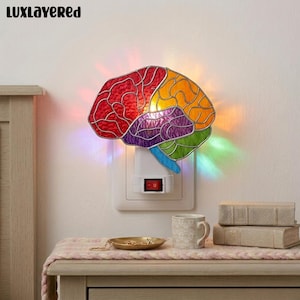 Puede incluir: Una luz de noche con forma de cerebro y colores vibrantes: rojo, naranja, morado, verde y azul. Se enchufa a una toma de corriente y tiene un interruptor de encendido/apagado rojo. La marca "LUXlayered" es visible en la parte superior izquierda.