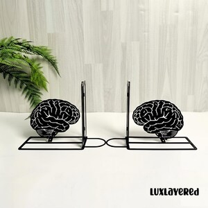 May include: Black metal bookends featuring a brain design. The bookends have a rectangular base and a vertical support. The brain design is a detailed, stylized illustration. The bookends are on a white surface.