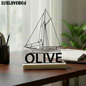 May include: Black metal sailboat sculpture with the word "OLIVE" at the base. The sailboat features a detailed outline of the boat, sails, and a person. The sculpture is mounted on a wooden base, creating a nautical theme.