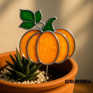 Könnte beinhalten: Ein Gartenstecker aus Buntglas in Form eines Kürbisses mit silbernem Metallumriss. Der Kürbis ist orange mit grünen Blättern und Stiel. Der Stecker befindet sich in einem Terrakotta-Topf mit einer Sukkulentenpflanze und kleinen weißen Steinen. Der Text "LUXLayeredRed" ist sichtbar.