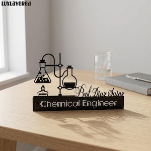 May include: Black desk sign featuring a stylized chemical engineering design with beakers, a flame, and the text "Chemical Engineer" and "Prof. Diar Sainz". The sign is on a wooden desk.