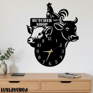 Può includere: Orologio da parete in metallo nero con un design a gallo, mucca e maiale, con la scritta "BUTCHER SHOP" sopra il quadrante. Le lancette sono dorate e i numeri bianchi.