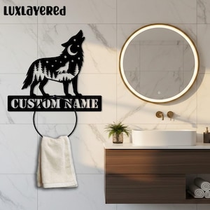Può includere: Portasciugamani in metallo nero con un design a lupo ululante. La silhouette del lupo include una foresta e stelle, con la scritta "CUSTOM NAME" sotto. Un asciugamano bianco è appeso al porta asciugamani circolare. Lo sfondo è un bagno.