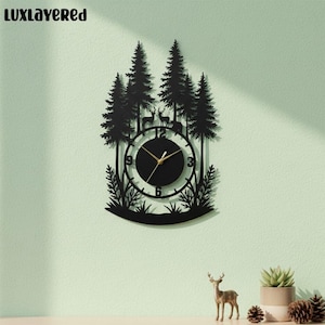 Puede incluir: Reloj de pared de metal negro con un diseño de escena de bosque. El reloj presenta una esfera circular con manecillas y números dorados, rodeada por una silueta de árboles, ciervos y follaje. El texto "LUXLAYERED" está en la parte superior izquierda.