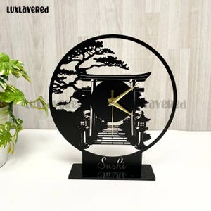 Könnte beinhalten: Schwarze Laser-Uhr mit japanischem Torii-Tor und Landschaftsdesign. Die Uhr hat goldfarbene Zeiger und das Wort "Sushi" an der Basis. Die Uhr steht auf einem schwarzen rechteckigen Sockel.