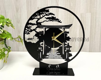 May include: Black laser-cut clock featuring a Japanese torii gate and landscape design. The clock has gold-colored hands and the word "Sushi" at the base. The clock is set on a black rectangular base.
