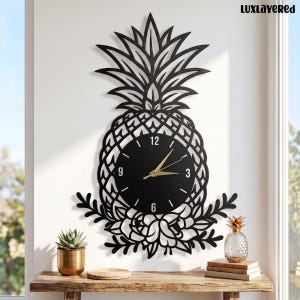 May include: A black wall clock shaped like a pineapple with a cutout leaf design at the top and floral accents at the base. The circular clock face is black with gold hands and white numerals at the 12, 3, 6, and 9 positions. The text LUXLAYERED is visible.