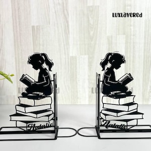 May include: Black metal bookends featuring a silhouette of a girl reading atop a stack of books. The bookends have a curved base and a vertical support. The name "Natalie" is written on the front of each bookend.