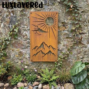 May include: A rectangular, rusty-brown metal wall art piece with a cut-out design of a sun, clouds, and mountains. The artwork is mounted on a stone wall with greenery and the text "luxlayered" at the top.