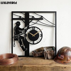 May include: Black metal wall clock featuring an electrician silhouette on a utility pole. The clock has a round face with gold hands and numbers. A brown leather tool belt and a hard hat with an American flag are also visible.