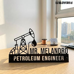 May include: A black metal desk sign featuring a silhouette of an oil pump jack, the text "MR. MELANDRO" in a bold sans-serif font, and the text "PETROLEUM ENGINEER" in a stencil-style font on a rectangular base with two circular screw holes.