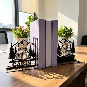 May include: A pair of black metal bookends featuring a laser-cut design of a log cabin, pine trees, and mountain peaks. The left bookend includes the word CABIN in capital letters at the base. The bookends support two light purple books.