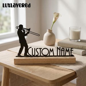 Könnte beinhalten: Eine schwarze Metallsilhouette einer Person, die Posaune spielt, neben dem Text "CUSTOM NAME" in schwarzer Schrift. Das gesamte Stück ist auf einem rechteckigen, hellen Holzsockel montiert und dient als dekoratives Tischobjekt.