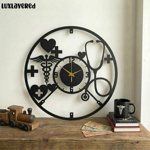 May include: Black metal wall clock with medical symbols, including a stethoscope, heart, and cross. The clock has gold hands and the text "LUXLAYERED" at the top. A black mug with a medical symbol is on a stack of books.