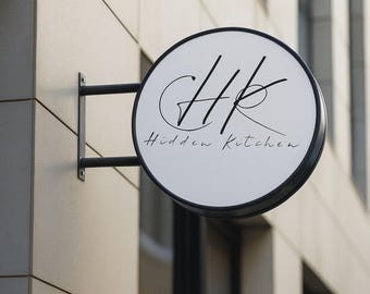 May include: A round, white sign with the text "HK Hidden Kitchen" in black script is mounted on a building. The sign is framed in black and attached to a black metal bracket. The background is a light-colored building facade.
