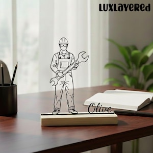 Puede incluir: Dibujo lineal en blanco y negro de un mecánico sosteniendo una llave inglesa, montado sobre una base de madera con el nombre "Olive". La imagen es un accesorio decorativo de escritorio. La marca "LUXLayered" está en la esquina superior derecha.