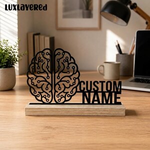 Könnte beinhalten: Ein schwarzer Ausschnitt eines menschlichen Gehirns mit integrierten Schaltkreismustern und dem Text "CUSTOM NAME", befestigt auf einem rechteckigen, hellen Holzsockel. Das Gehirndesign zeigt zwei symmetrische Hälften mit Linien.