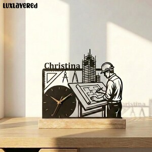 May include: A black metal desk clock featuring a silhouette of an engineer wearing a hard hat, working at a drafting table with blueprints. The design includes a skyscraper, a triangle, a compass, and the name Christina on a wooden base.