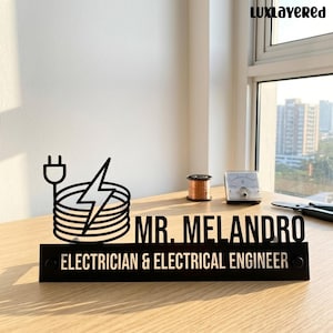 Puede incluir: Un letrero de escritorio negro con un gráfico de un cable enrollado, un enchufe y un rayo. El texto dice "MR. MELANDRO" sobre "ELECTRICIAN & ELECTRICAL ENGINEER". El letrero está colocado sobre una superficie de madera cerca de una bobina de cable de cobre y un medidor.