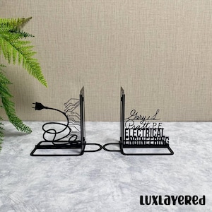 Puede incluir: Soportes para libros de metal negro con un diseño de enchufe eléctrico y placa de circuito. Un sujetalibros presenta el texto "Gary L Pratt P.E. ELECTRICAL ENGINEERING". Los sujetalibros están sobre una superficie gris.