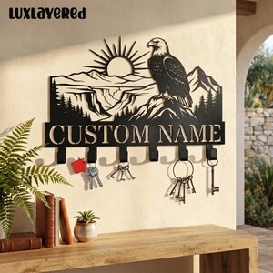 Pode incluir: Um suporte de chaves de parede em metal preto com um design cortado a laser de uma águia pousada numa montanha com um sol nascente. A peça inclui o texto CUSTOM NAME e seis ganchos, com várias chaves de metal penduradas neles.