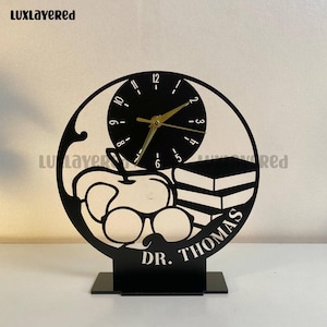 May include: Black laser-cut desk clock featuring a clock face, an apple, glasses, and books. The clock has gold hands and the name "DR. THOMAS" is displayed on the base. The clock is a decorative item.