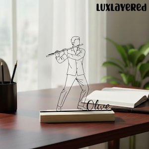 May include: A black and white line drawing of a person playing a flute, mounted on a wooden base. The name "Olive" is written in cursive on the base. The logo "LUXLayered" is in the top right corner. The item is on a dark brown desk.