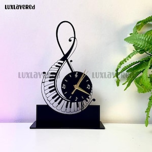 May include: Black decorative clock shaped as a treble clef and piano keys. The clock face is black with gold hands and musical note symbols. The base is a black rectangular shape. The brand name "LUXLAYERED" is printed on the top left and bottom right.