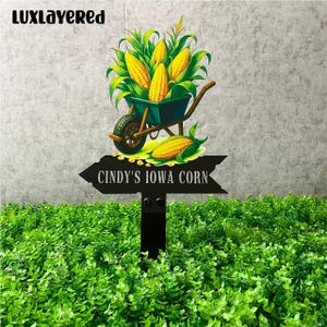 Könnte beinhalten: Ein schwarzes Metall-Gartenschild mit dem Text "Cindy's Iowa Corn". Das Schild zeigt eine Schubkarre voller Mais und Maiskolben. Das Schild steht in einem Beet mit grünen Pflanzen.