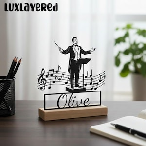 Puede incluir: Figura de director de orquesta negra cortada con láser con notas musicales y el nombre "Olive" sobre una base de madera. El director lleva un esmoquin y sostiene una batuta. El logotipo de Luxlayered está en la esquina superior izquierda.