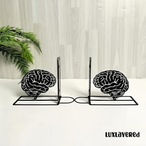 May include: Black metal bookends featuring a brain design. The bookends have a rectangular base and a vertical support. The brain design is a detailed, stylized illustration. The bookends are on a white surface.