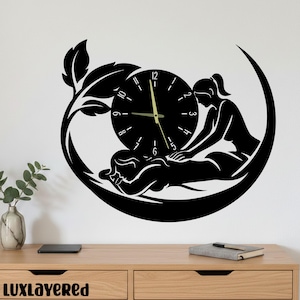 May include: Black wall clock featuring a spa scene with a person receiving a massage. The clock face has a minimalist design with numbers and hands. The clock is surrounded by a crescent shape and leaf accents.