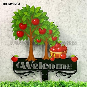 Puede incluir: Un letrero de bienvenida de metal negro con un diseño decorativo, con dos manzanos con manzanas rojas y una cesta de manzanas. El letrero se coloca frente a un área verde y cubierta de hierba.