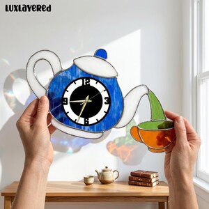 May include: A stained glass wall clock shaped like a teapot in blue and white, pouring a green liquid into an orange teacup. The clock face is black and white with gold hands. The text LUXLAYERED is visible in the top left corner of the image.