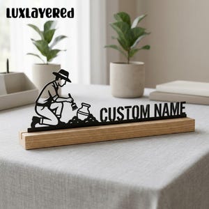 Peut inclure: Une silhouette en métal noir représentant un archéologue agenouillé brossant la terre d'un vase enterré, fixée sur une base rectangulaire en bois clair. Le texte CUSTOM NAME est découpé en métal noir à côté, avec le texte LUXLAYERED au-dessus.
