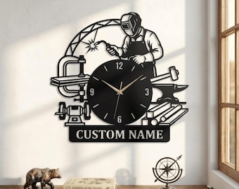 Puede incluir: Un reloj de pared de metal negro con la silueta de un soldador trabajando, una abrazadera, un martillo, un yunque y vigas de metal. La esfera tiene manecillas doradas y los números 3, 6, 9 y 12. Una placa inferior muestra el texto CUSTOM NAME.