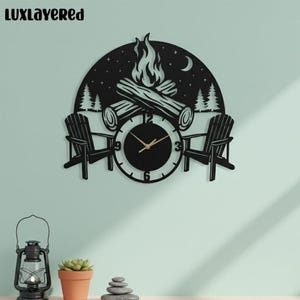 May include: Black metal wall clock featuring a campfire scene with two Adirondack chairs. The clock face is black with gold hands, and the design includes a crescent moon, trees, and the text "LUXLAYERED".