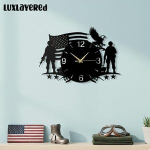 Pode incluir: Relógio de parede preto com um design de silhueta apresentando uma bandeira americana, uma águia e soldados. O relógio tem ponteiros dourados e os números 12, 3, 6 e 9 são visíveis. A marca "LUXLAYERED" está no topo.