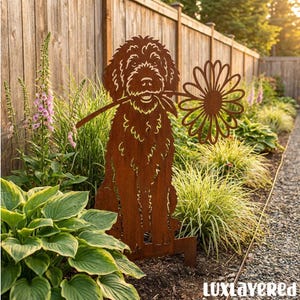Op de afbeelding: Een roestkleurige metalen tuinsteker met een uitgesneden silhouet van een ruige hond die een grote margriet in de bek houdt. Het platte, decoratieve figuur is ontworpen om in de grond te worden gestoken als tuinornament.