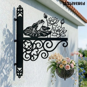 Könnte beinhalten: Schwarzer Metall-Hängepflanzenhalter mit einem Frosch- und Lotusblumen-Design. Der Halter trägt einen Hängekorb mit rosa und gelben Blumen und Grünzeug. Der Halter ist an einer weißen Wand montiert.