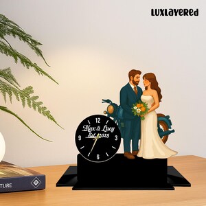 Könnte beinhalten: Eine schwarze Uhr mit weißen Ziffern und Zeigern, die ein Paar und ein Motorrad zeigt. Die Uhr zeigt "Max & Lucy Est. 2025". Das Paar steht vor einem blauen Motorrad, die Frau in einem weißen Kleid und der Mann in einem blauen Anzug.
