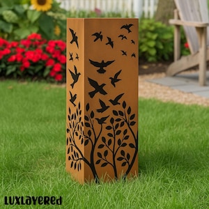 May include: A rectangular, brown metal garden sculpture with a design of black birds in flight and stylized trees with leaves. The sculpture is placed on a green lawn, with red flowers and a wooden chair in the background.