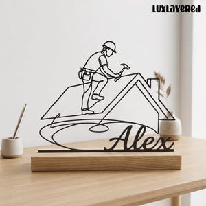 Könnte beinhalten: Eine schwarze Metalldrahtskulptur, die einen Bauarbeiter mit einem Hammer auf einem Dach darstellt, montiert auf einem rechteckigen Holzsockel. Der Name Alex ist unten in Schreibschrift geschrieben. Der Text LUXLAYERED erscheint oben rechts.