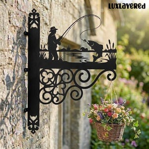 May include: Black metal hanging sign featuring a fisherman silhouette, fishing rod, and a leaping fish. The sign is mounted on a brick wall and supports a hanging basket filled with colorful flowers. The text "LUXLAYERED" is in the upper right corner.