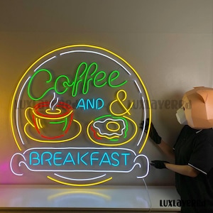 May include: A neon sign with the words "Coffee AND BREAKFAST" in a circular design. The sign features a coffee cup, steam, and a donut. The sign has a yellow, white, green, red, and blue color scheme.